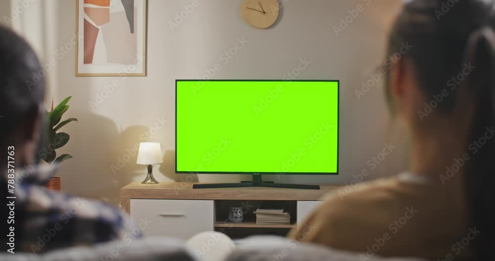 Rear view of multiethnic couple sitting at sofa. Young couple sitting in front of TV set with chroma key screen. Man and woman looking at green mock up screen. Multucultural couple sitting at couch. Rear view of multiethnic couple sitting at sofa. Young couple sitting in front of TV set with chroma key screen. Man and woman looking at green mock up screen. Multucultural couple sitting at couch.