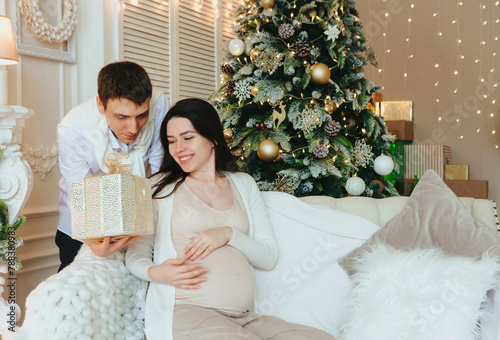 Husband giving Christmas present to happy surprised pregnant wife while sitting beside xmas tree © Dasha Petrenko