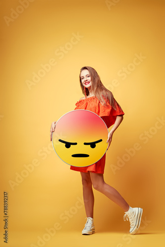 Social network concept. Happy young woman holding yellow angry emoji face. Colorful studio portrait. © luengo_ua