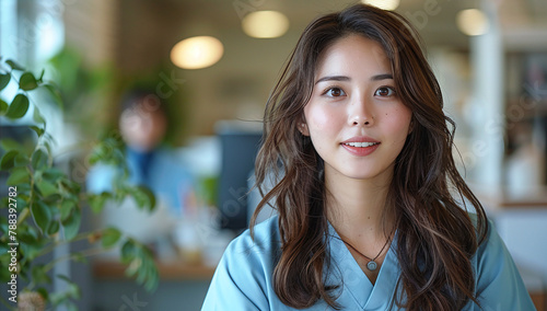 Portrait of a young Asian woman with long wavy hair, wearing a blue scrub, smiling in a bright office environment.