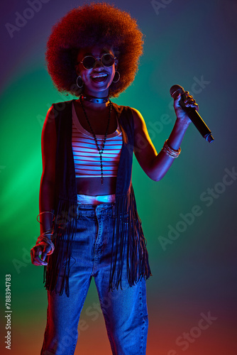 Afro-haired musician in sunglasses, gripping microphone, intense colored in neon light against gradient background. Concept of art, music, hobby, classical music and modern lifestyle. Ad