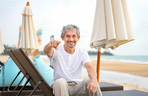 playful elderly man sitting on beach chair,smiling and pointing to camera,happy senior male enjoy holiday time on the beach © Verin