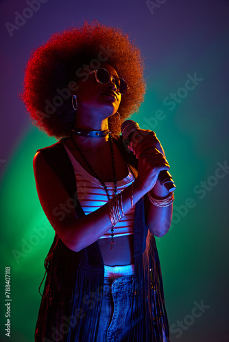 Young vocalist with iconic 70s afro, singing, dressed in black vest and beaded necklaces in neon light against gradient background. Concept of art, music, hobby, classical music and modern lifestyle.