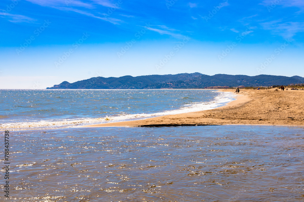 En la Playa de la Estartit, Costa Brava, la luz del sol de primavera danza sobre las aguas y la arena, con montañas lejanas como telón de fondo.