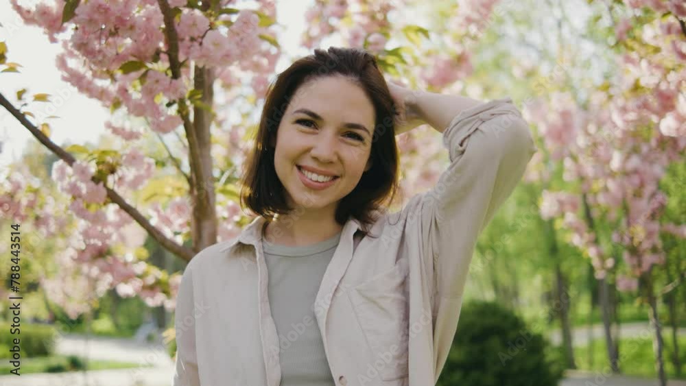 Portrait of a beautiful young woman. A girl walks in the park among pink blossoming sakura or cherry trees. Female smiles and looks at the camera against the background of pink spring trees on sunset Portrait of a beautiful young woman. A girl walks in the park among pink blossoming sakura or cherry trees. Female smiles and looks at the camera against the background of pink spring trees on sunset