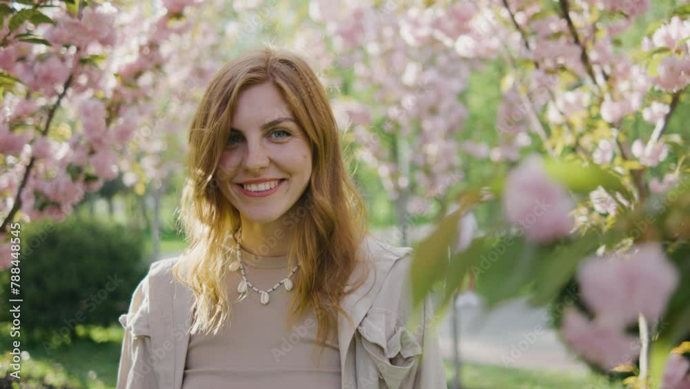 Portrait of a beautiful young red-haired woman. A girl walks in the park among pink blossoming sakura or cherry trees. Female smiles and looks at the camera against the background of pink spring trees Portrait of a beautiful young red-haired woman. A girl walks in the park among pink blossoming sakura or cherry trees. Female smiles and looks at the camera against the background of pink spring trees