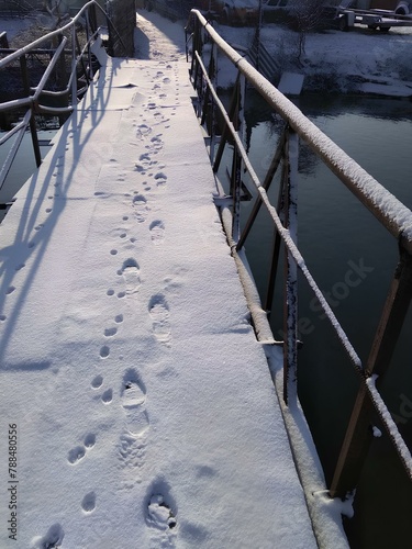 A narrow bridge covered with snow, with human footprints in the snow leading across the bridge. Water is visible on both sides. The sun illuminates the scene, creating a bright and wintry day photo