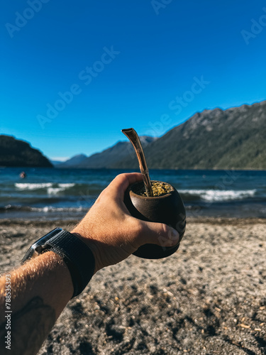 A person holding a small cup with a plant in it on a beach. The beach is near a body of water and the sky is blue