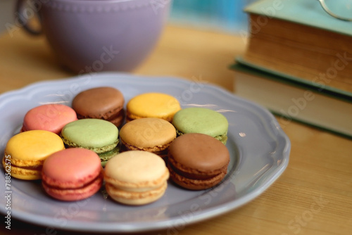 Purple plate filled with pastel macarons, cup of tea or coffee, vintage books and reading glasses on the table. Colorful bookcase in the background. Selective focus.
