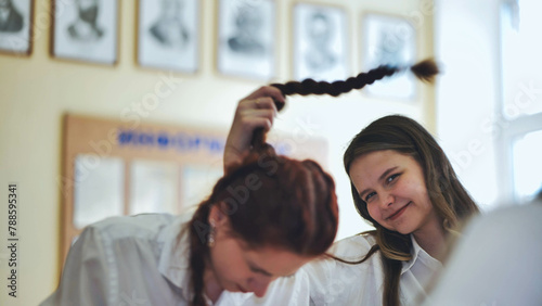 Fun schoolgirls in the classroom. Girl playing with her friend s pigtail.
