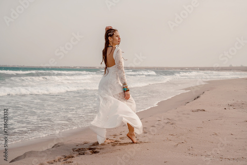 Model in boho style in a white long dress and silver jewelry on the beach. Her hair is braided  and there are many bracelets on her arms.