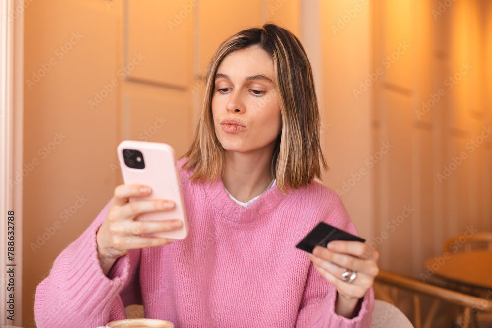 Calm young woman wear pink knit sweater clothes with credit card in hand using mobile phone paying for purchase online relaxing sitting at table in cafe. Serious blonde girl. Calm young woman wear pink knit sweater clothes with credit card in hand using mobile phone paying for purchase online relaxing sitting at table in cafe. Serious blonde girl.