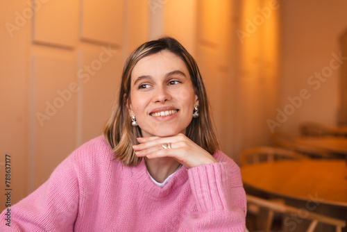 Happy young woman sitting on chair at cafe and looking at side. Portrait of comfortable woman in winter clothes relaxing on armchair. Portrait of beautiful girl smiling and relaxing during autumn.