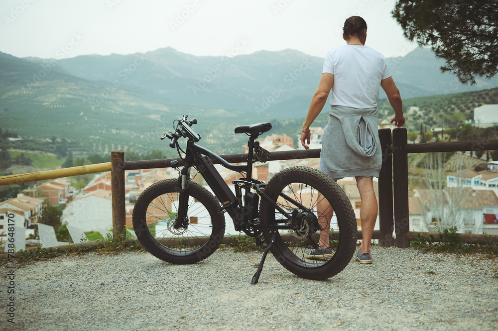 © Taras Grebinets - View from the back of a young man cyclist, standing near his electric bike, having a break after biking in mountains. People. Active healthy lifestyle. E-bike as sustainable, eco friendly transport © Taras Grebinets - View from the back of a young man cyclist, standing near his electric bike, having a break after biking in mountains. People. Active healthy lifestyle. E-bike as sustainable, eco friendly transport
