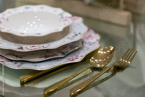 Stack of porcelain dishes with golden cutlery on a glass table. Below the bowl are plates of increasing sizes with floral designs. A golden spoon and fork are placed beside the stacked dishes.