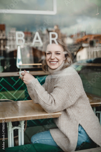 Smiling Woman Enjoying Coffee in Cafe. A cheerful woman sits at a cafe table, holding a cup of coffee with a visible bar sign in the background. Warmth and relaxation exude from the cozy atmosphere