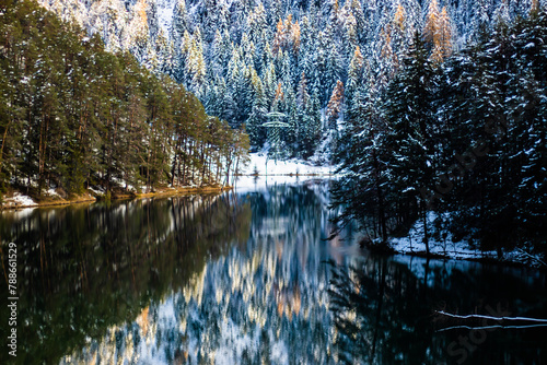 Paisagem de um lago na floresta, com árvores e neve, onde se vê o reflexo de tudo na água