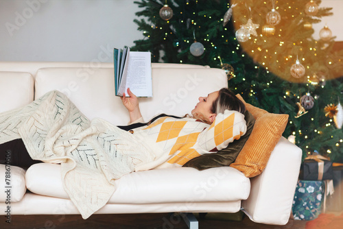 Relaxed woman reading book while lying on sofa in living room with decorated Christmas tree photo