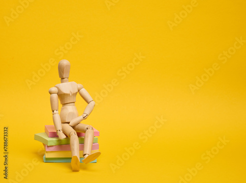 A wooden puppet doll sits on a stack of paper multi-colored stickers on a yellow background