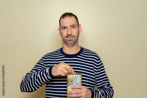 A Hispanic man with a beard wearing a striped sweater putting a euro coin into a metal piggy bank, trying to save so he can travel on vacation. Isolated on beige studio background.