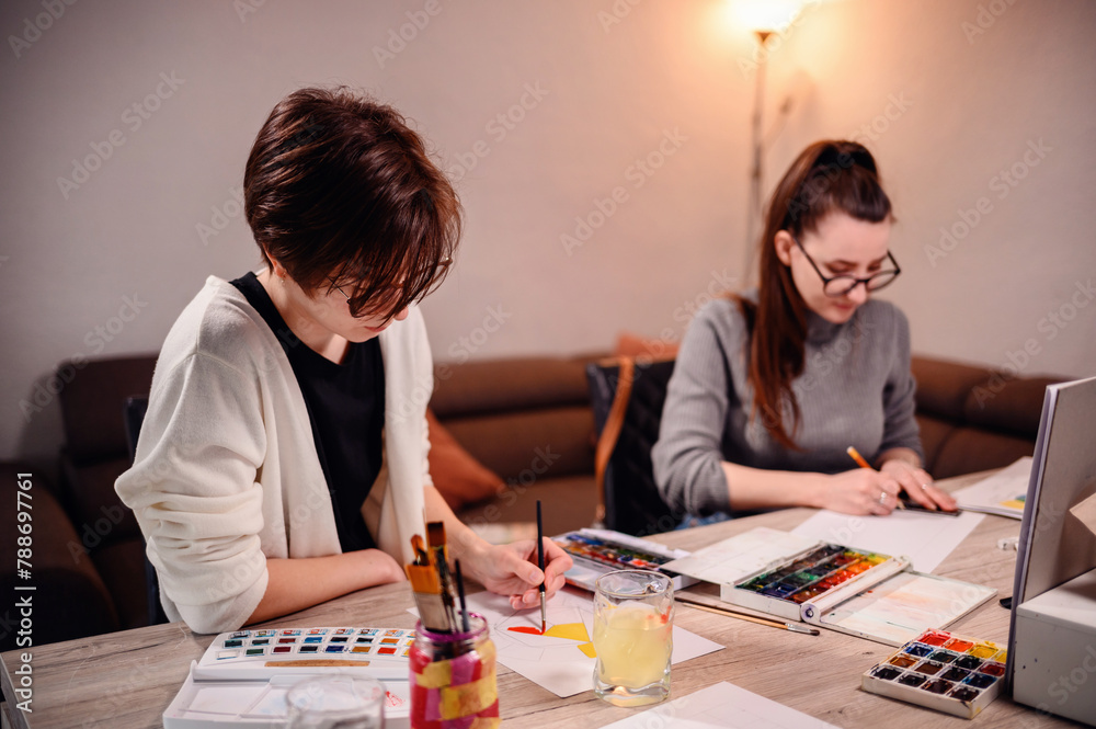 Two concentrated women involved in an artistic project, with one painting and the other measuring, surrounded by creative tools