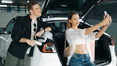 Young couple taking selfie with EV electric car charging battery at shop center parking lot n city downtown as green urban sustainable lifestyle by rechargeable energy of EV vehicle innards