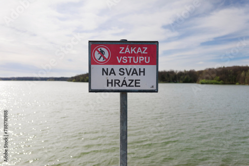 Warning sign with text - Do not enter on the dam embankment (translated from Czech). Prohibition, bad and interdiction. Water reservoir in the background. Shallow focus.