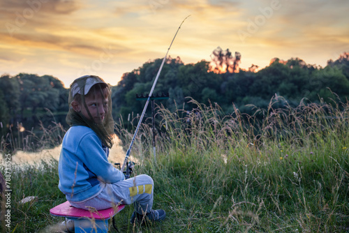 Young boy sitting with fishing rod on the edge of a high riverbank at sunset with vibrant colors on the sky © skumer