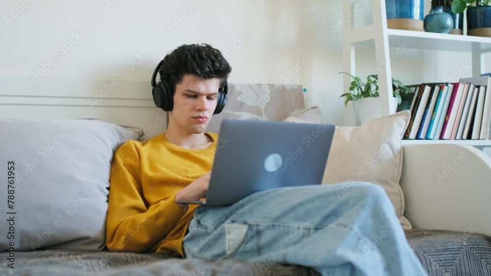 Young guy college student in headphones typing on laptop sitting on couch at home Young guy college student in headphones typing on laptop sitting on couch at home