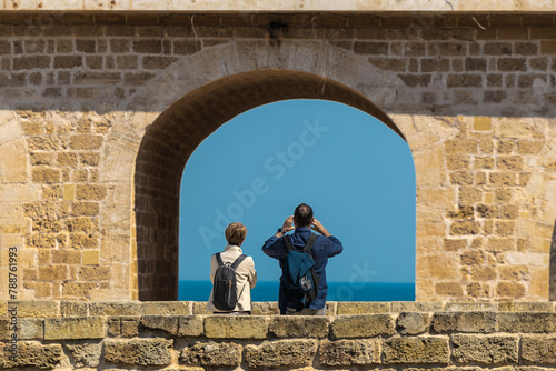 A tourist couple is photographing beach and sea from the bridge in Polignano a Mare, popular spot for tourists and locals. Warm spring setting, stone wall