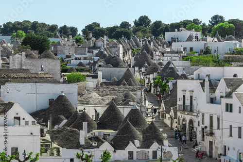  Beautiful stone Trulli houses with narrow streets in village of Alberobello. Picturesque village on a hill in Apulia, southern italy. Green trees and white houses with stone roofing