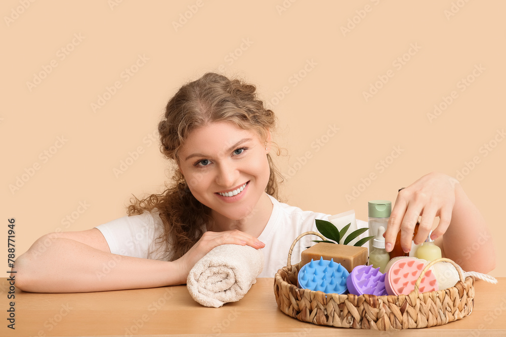 Beautiful young woman with hair scalp massagers and cosmetic products at table against beige background Beautiful young woman with hair scalp massagers and cosmetic products at table against beige background