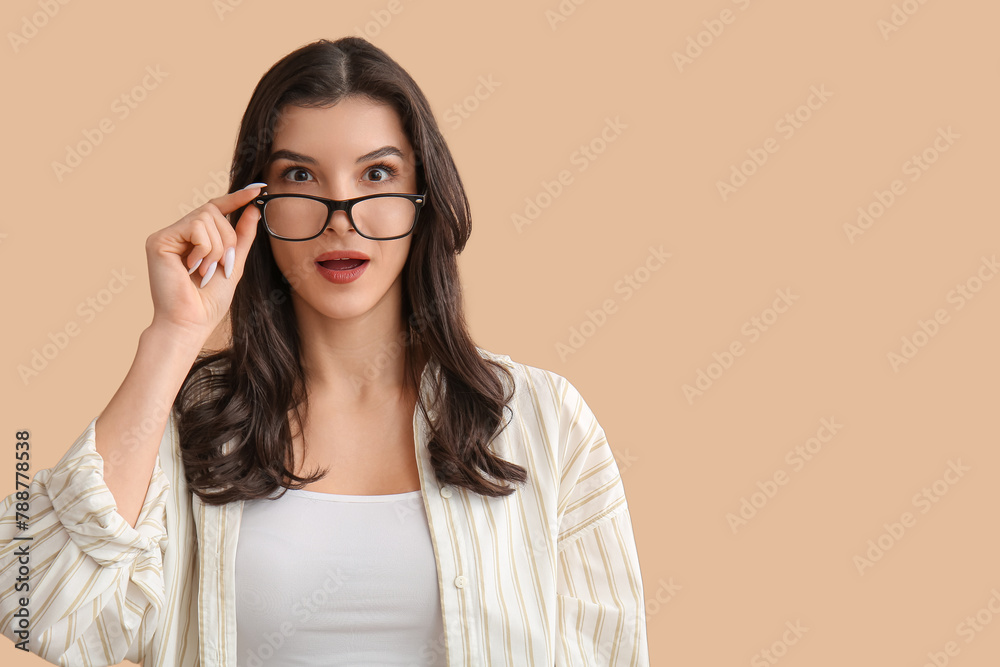 Surprised young woman putting on eyeglasses on beige background