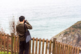 Close-up shot of a Japanese tourist with his back to a professional camera taking a picture of the landscape of the touristy Basque coastline