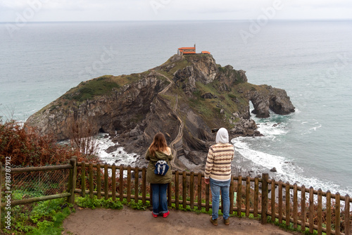 Couple of tourists with their backs to San Juan de Gaztelugatxe taking pictures of the hermitage surrounded by the sea on the touristy Basque coast.