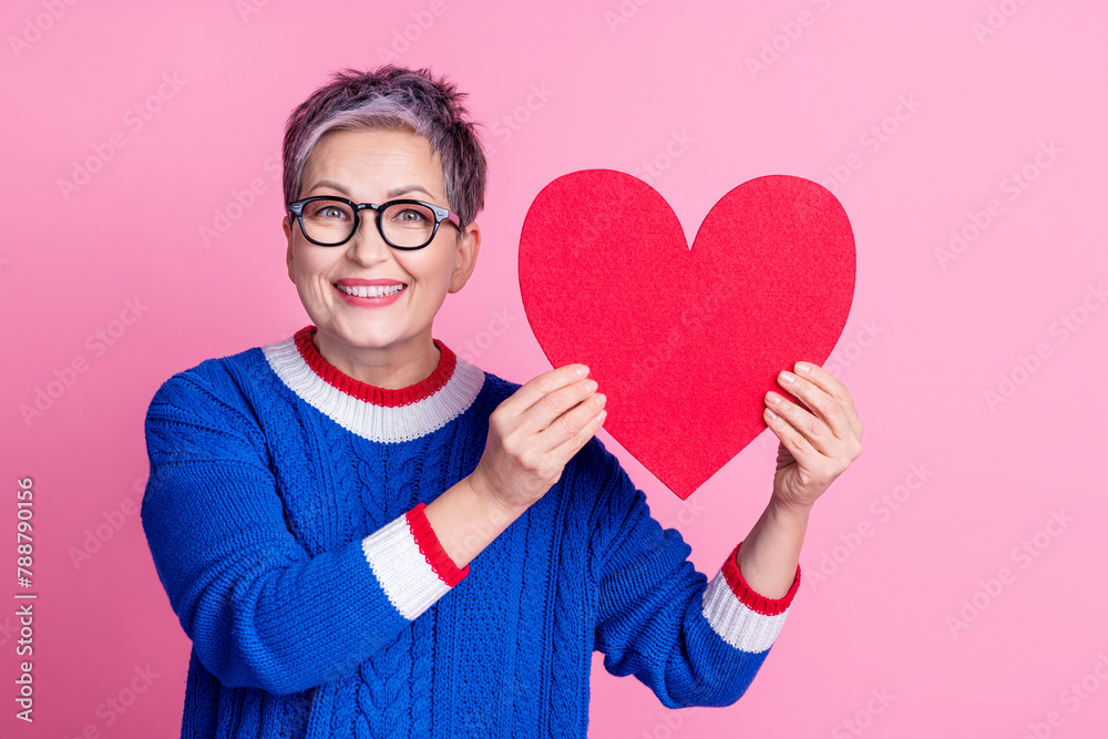 Photo of cute lovely senior woman wear stylish blue clothes hands hold big large red paper card isolated on pink color background Photo of cute lovely senior woman wear stylish blue clothes hands hold big large red paper card isolated on pink color background