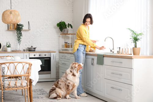 Pretty young woman washing dishes with cute Australian Shepherd dog in kitchen