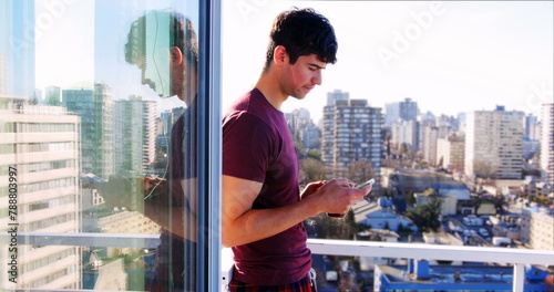 Man in purple shirt standing on balcony using smartphone, cityscape in background