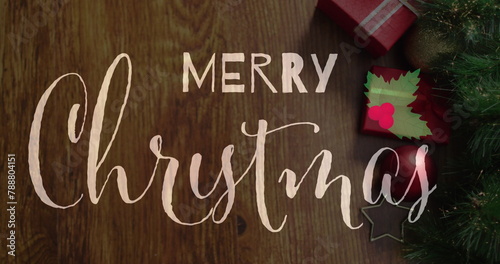 Merry Christmas written in cursive on wooden surface, surrounded by holiday decor