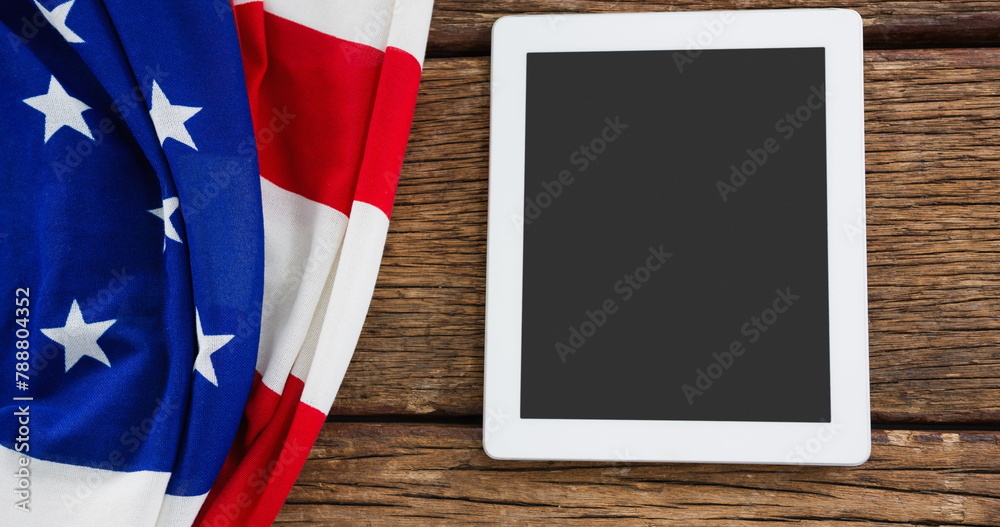 Fototapeta premium American flag draping next to white tablet on a wooden surface