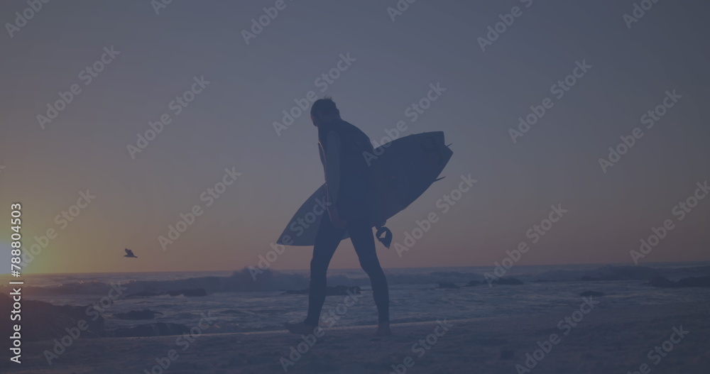 Obraz premium Caucasian young adult walking on beach, holding surfboard