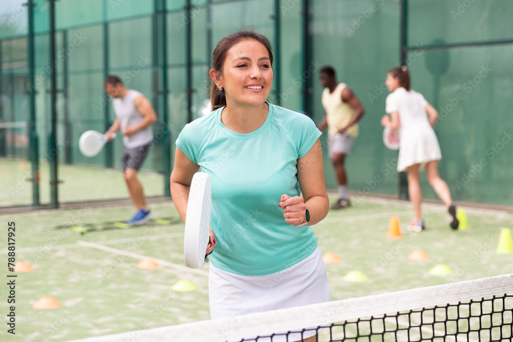 Portrait of cheerful woman paddle tennis player performing strokes technique at group training at court