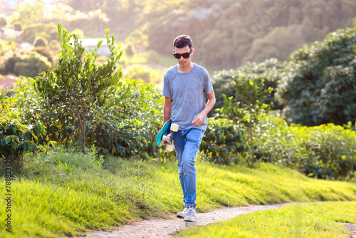 Man walks outdoors, carrying his longboard skate, seeking a spot to ride, with a non-urban landscape in the background. Promotes outdoor activities and exploration © Marcio