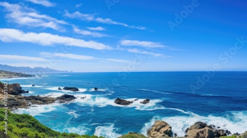 Beautiful seascape with blue ocean waves crashing on rocky coast. Clear blue sky with white clouds in background.