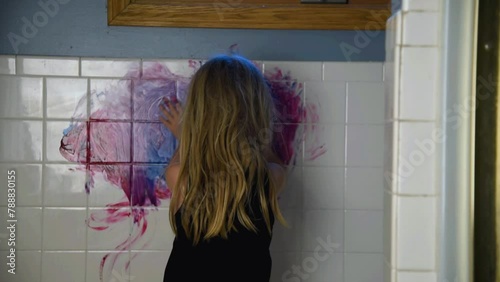 A creative little child is painting on a bathroom wall with her hands making a mess for a funny parenting discipline concept.