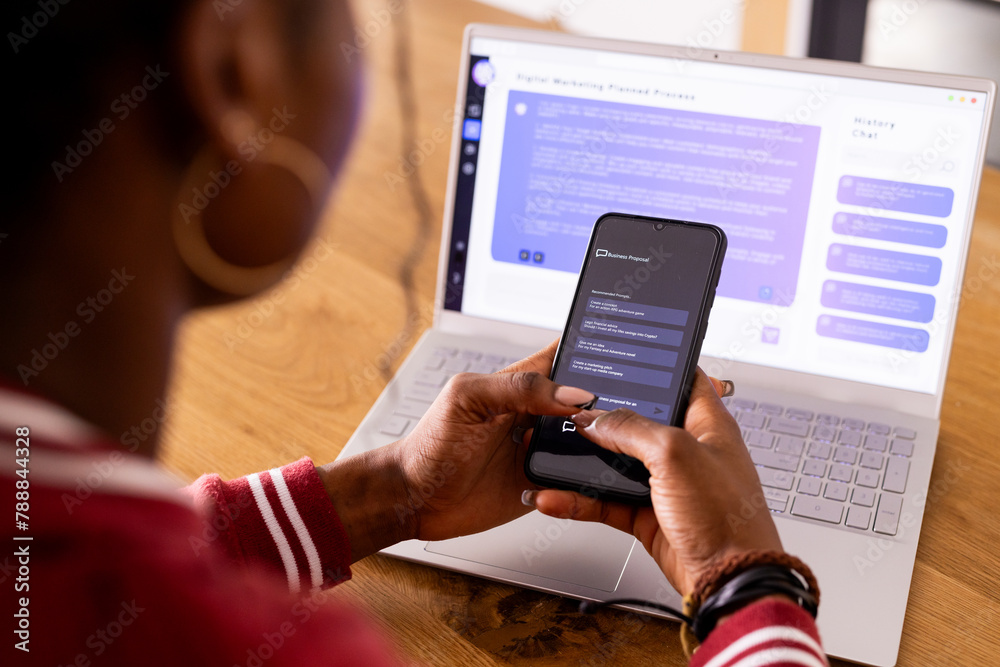 © Wavebreak Media - African American young woman indoors with phone, laptop showing AI Chat Bot Screen at home