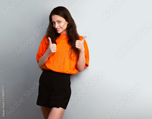 Happy toothy smiling beautiful woman showing thumb up sign by two hands in bright orange t-shirt and black skirt on blue studio wall background