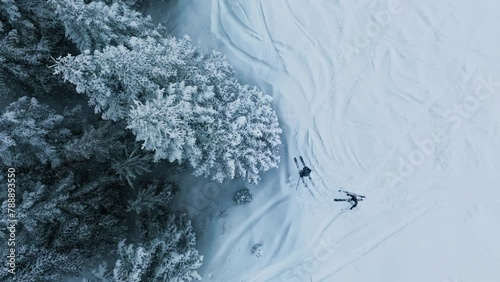Slow motion overhead shot of skiers at Stevens Pass ski resort with snowy mountain forest. Top down aerial ski hill on winter day during snowfall in Washington.Two skiers skiing on snow slope in pines photo
