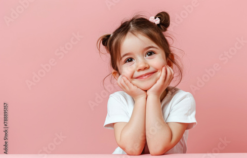 Cute little girl smiling and looking up, holding her chin with both hands on pink background © Kien