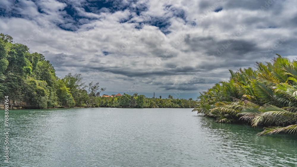 A calm turquoise river in the jungle. There are thickets of palm trees and tropical vegetation on the shores. Clouds in the blue sky. Philippines. Bohol. Loboc river.